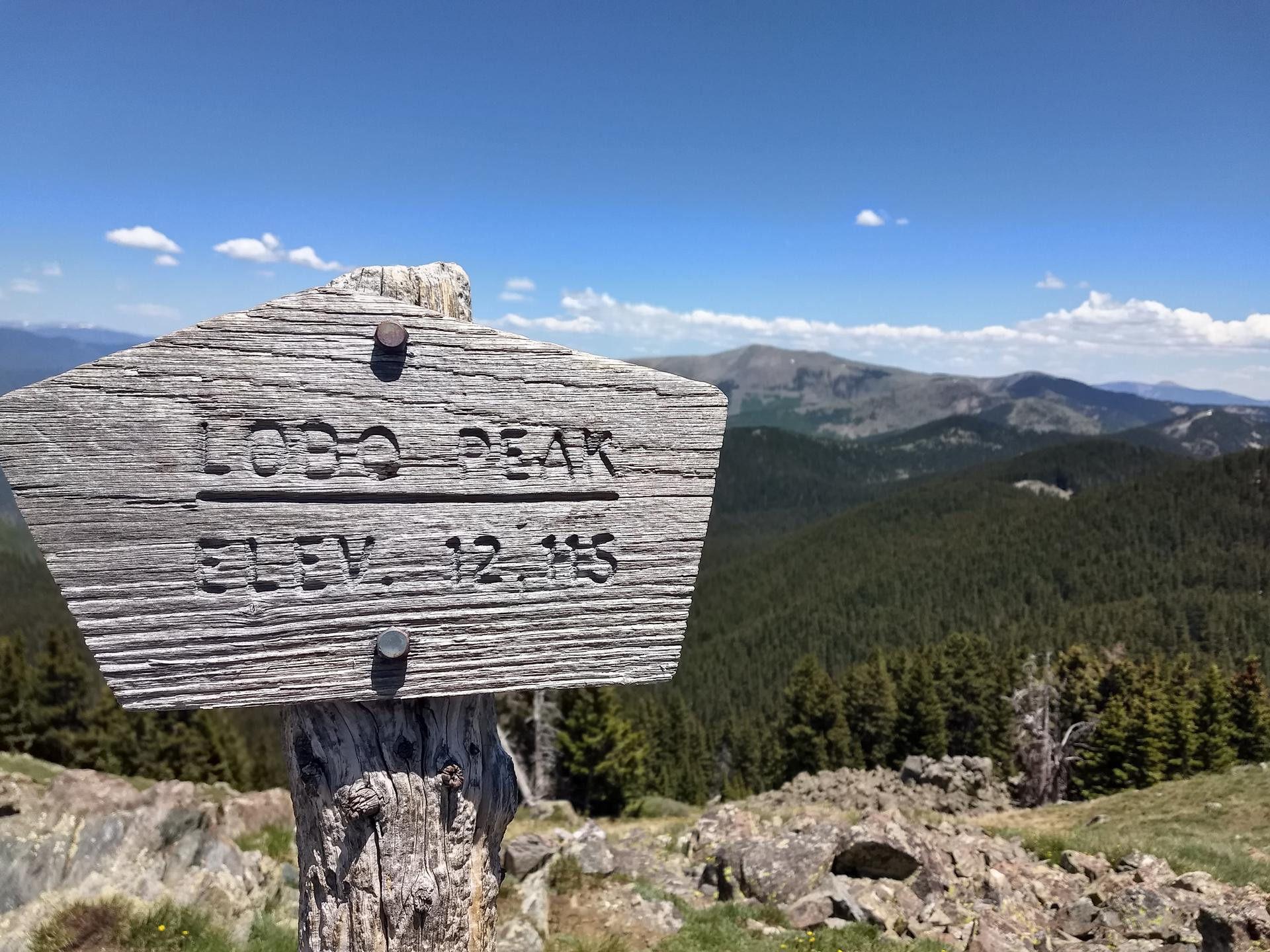 Weathered wooden sign on a mountain peak with forests under a bright blue sky.