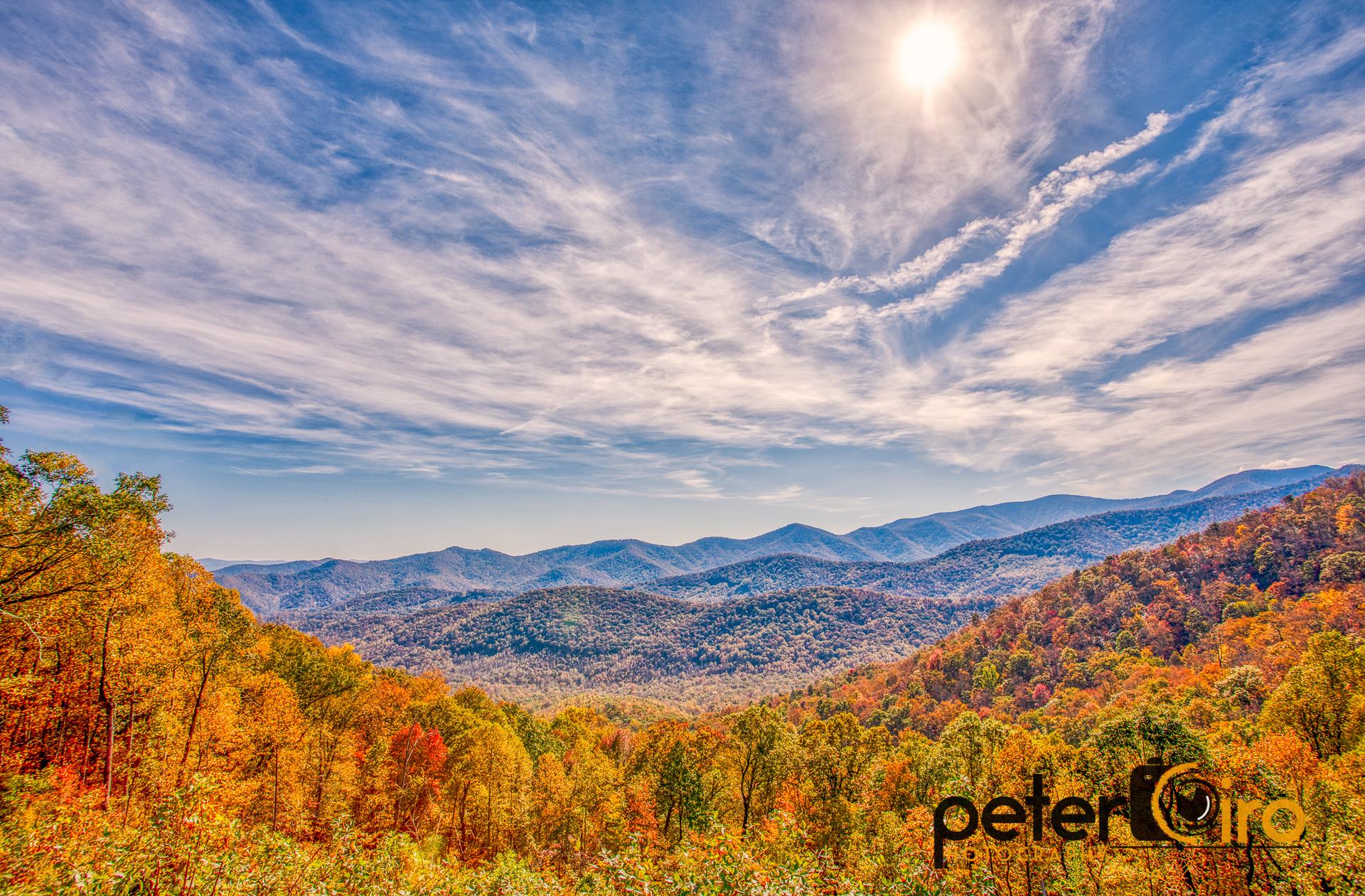 Vast mountain landscape with vibrant autumn trees under a bright, partly cloudy sky.