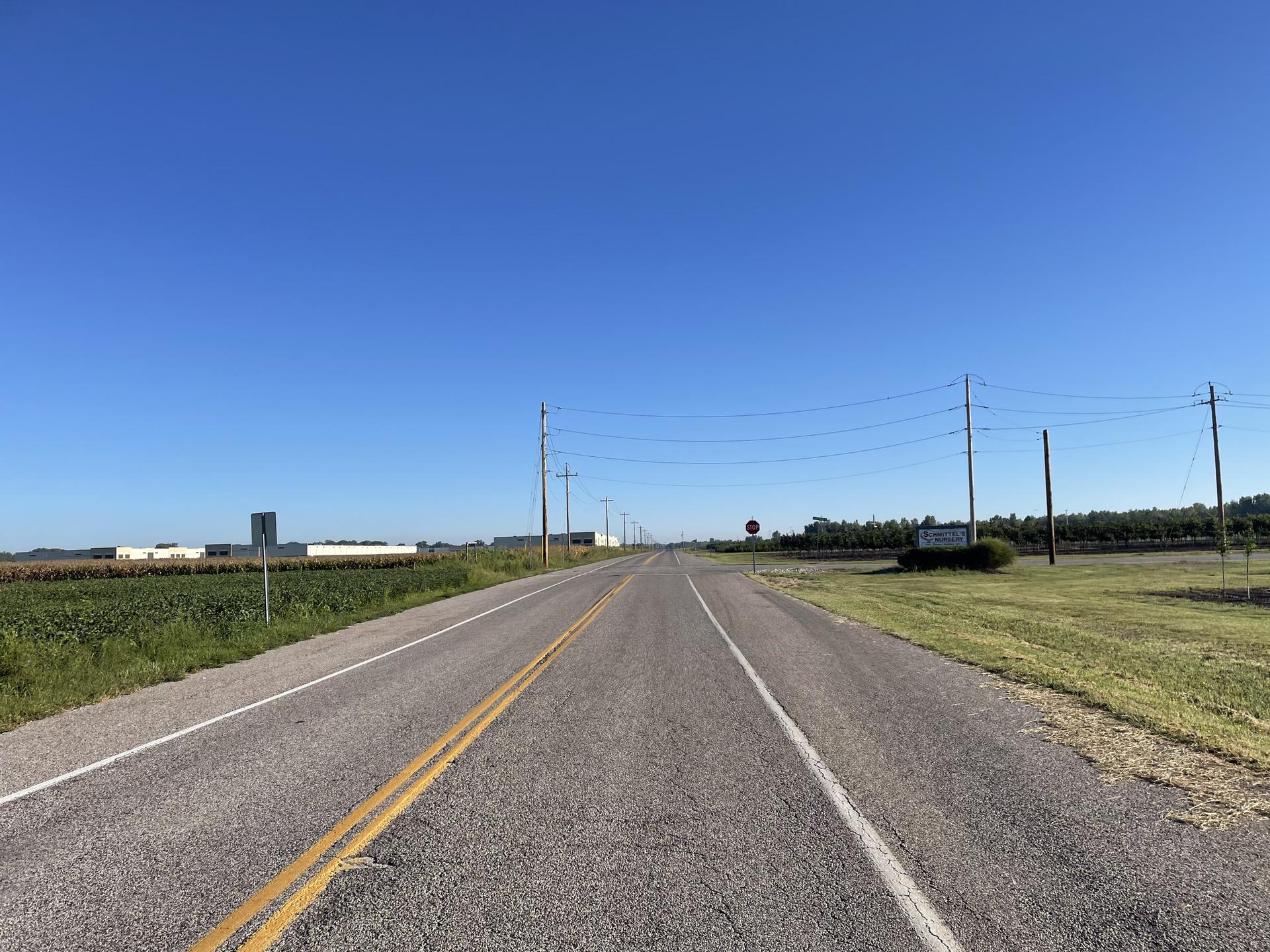 A long, empty rural road stretches through fields under a clear sky.