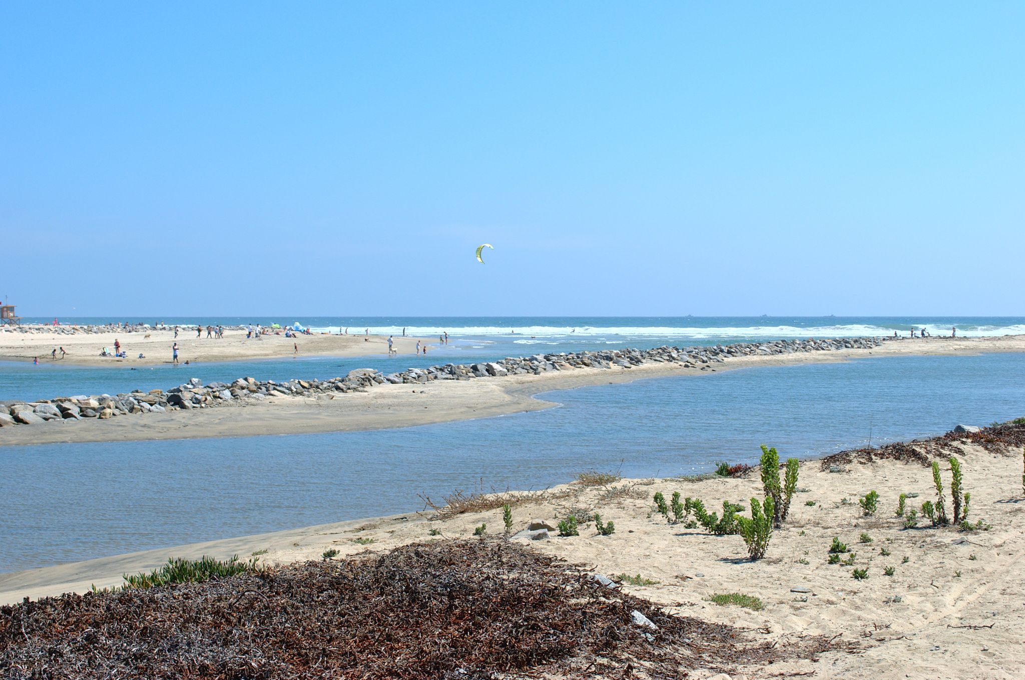Sunny beach scene with a water channel, rock barriers, and distant ocean. - Least Tern Natural Preserve – Santa Ana River Trail loop from Orange Coast College