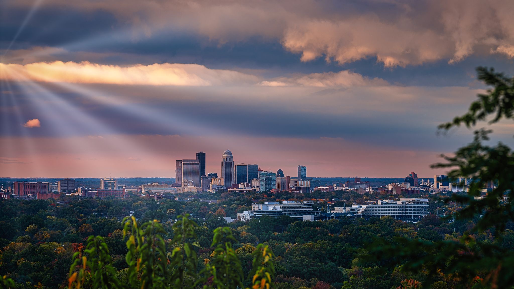 City skyline under dramatic sun rays breaking through clouds over green trees. - Iroquois Park Scenic Loop