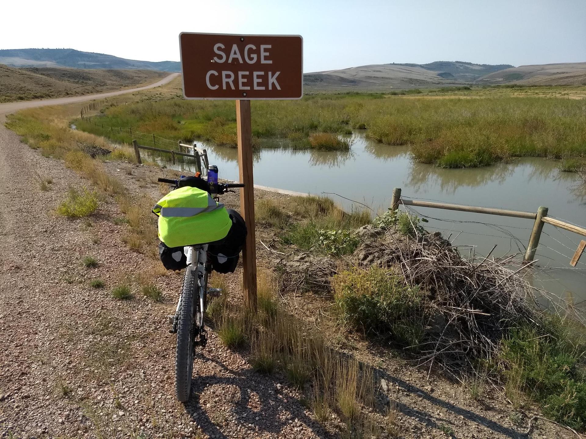 A bicycle with bright panniers rests by a sign next to murky water and reeds.