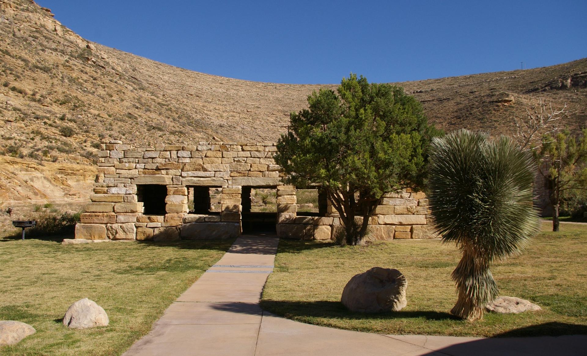 Stone building with open doorways on a green lawn, arid hills in background.