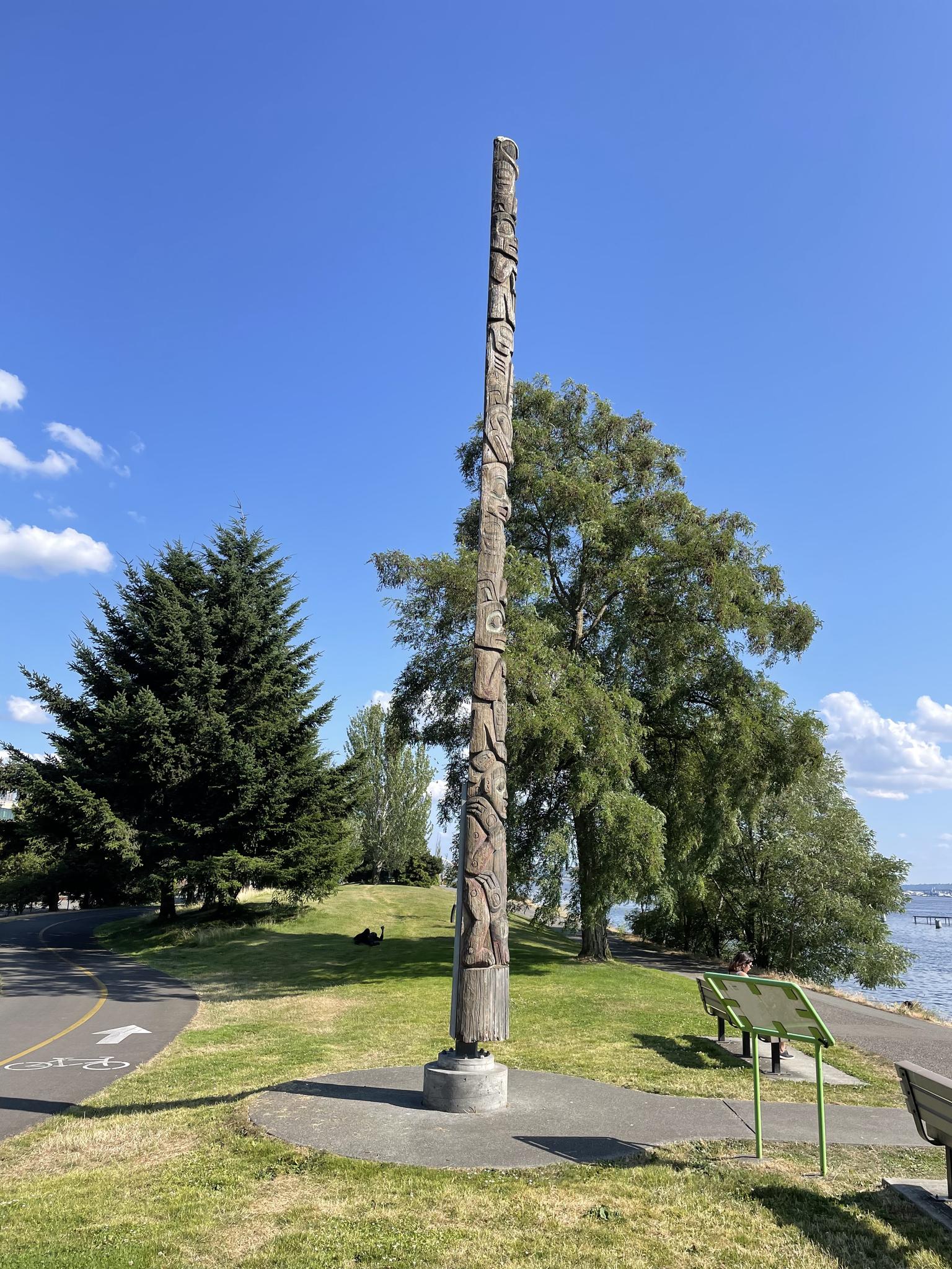 Tall carved wooden totem pole in grassy park with trees under clear blue sky.