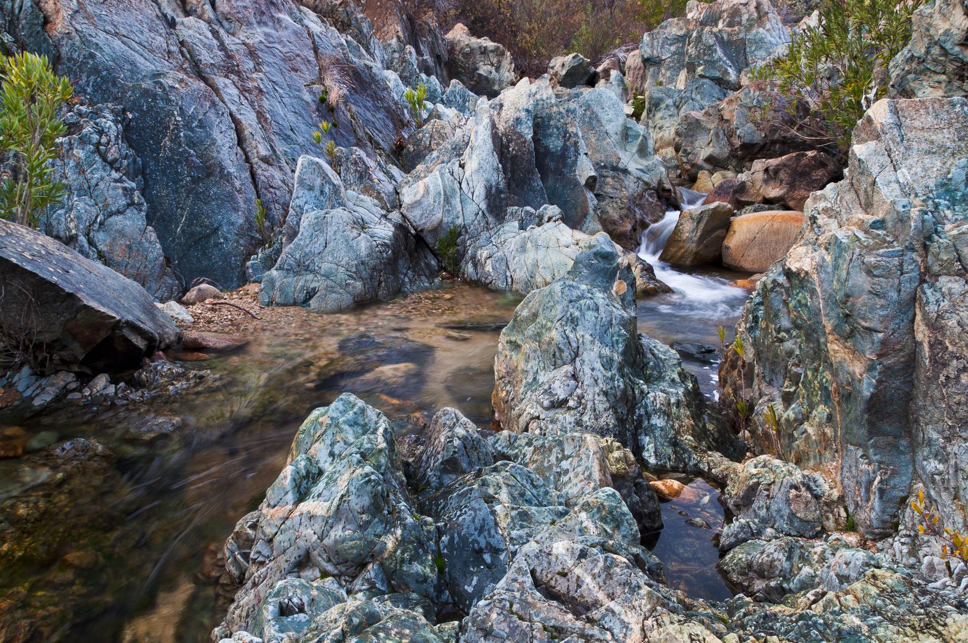 A clear stream flows through a rugged, rocky landscape with green foliage.