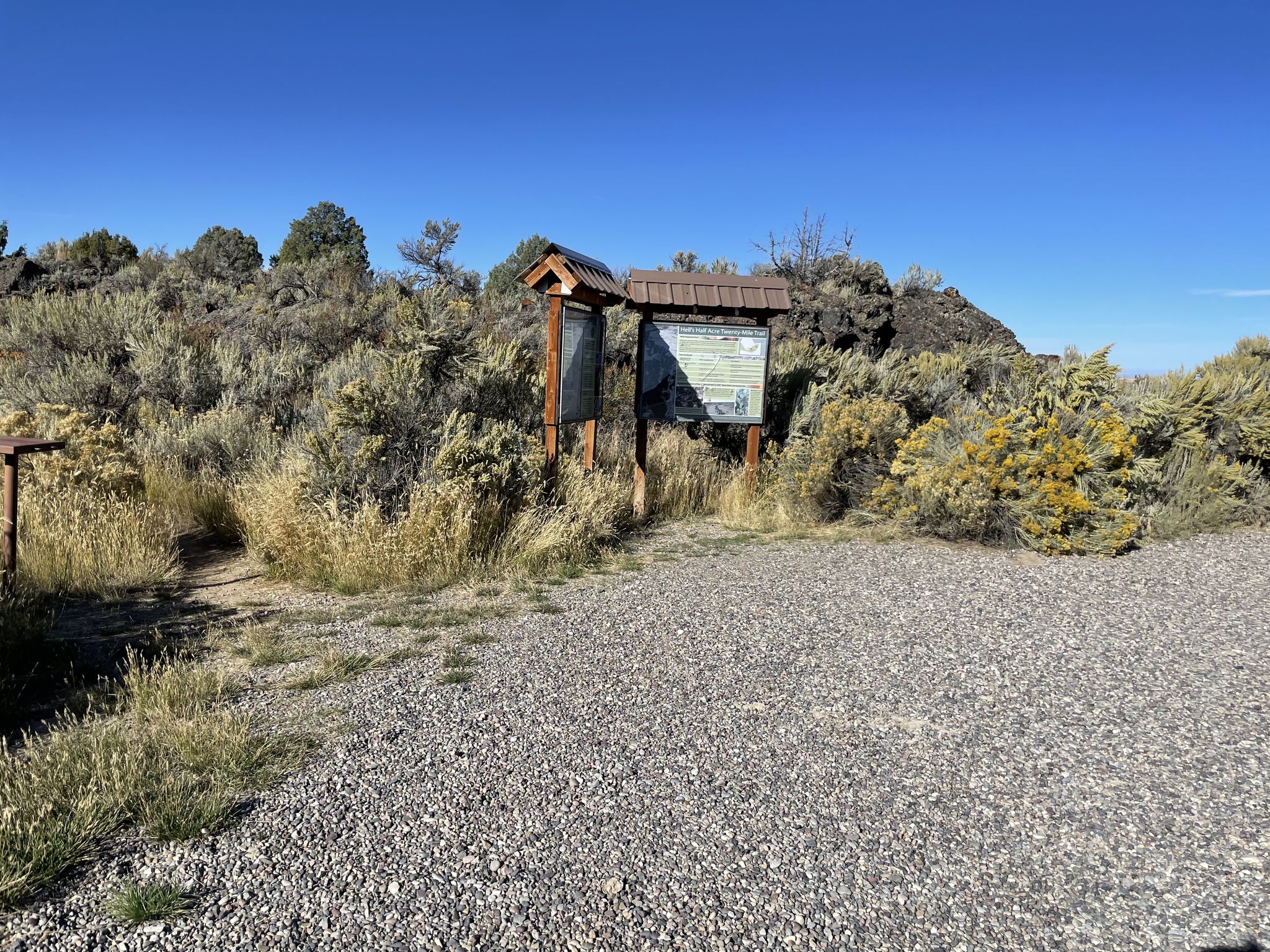 Wooden information kiosk on a gravel path, surrounded by dry bushes, under clear sky. - Lava Loop Trail