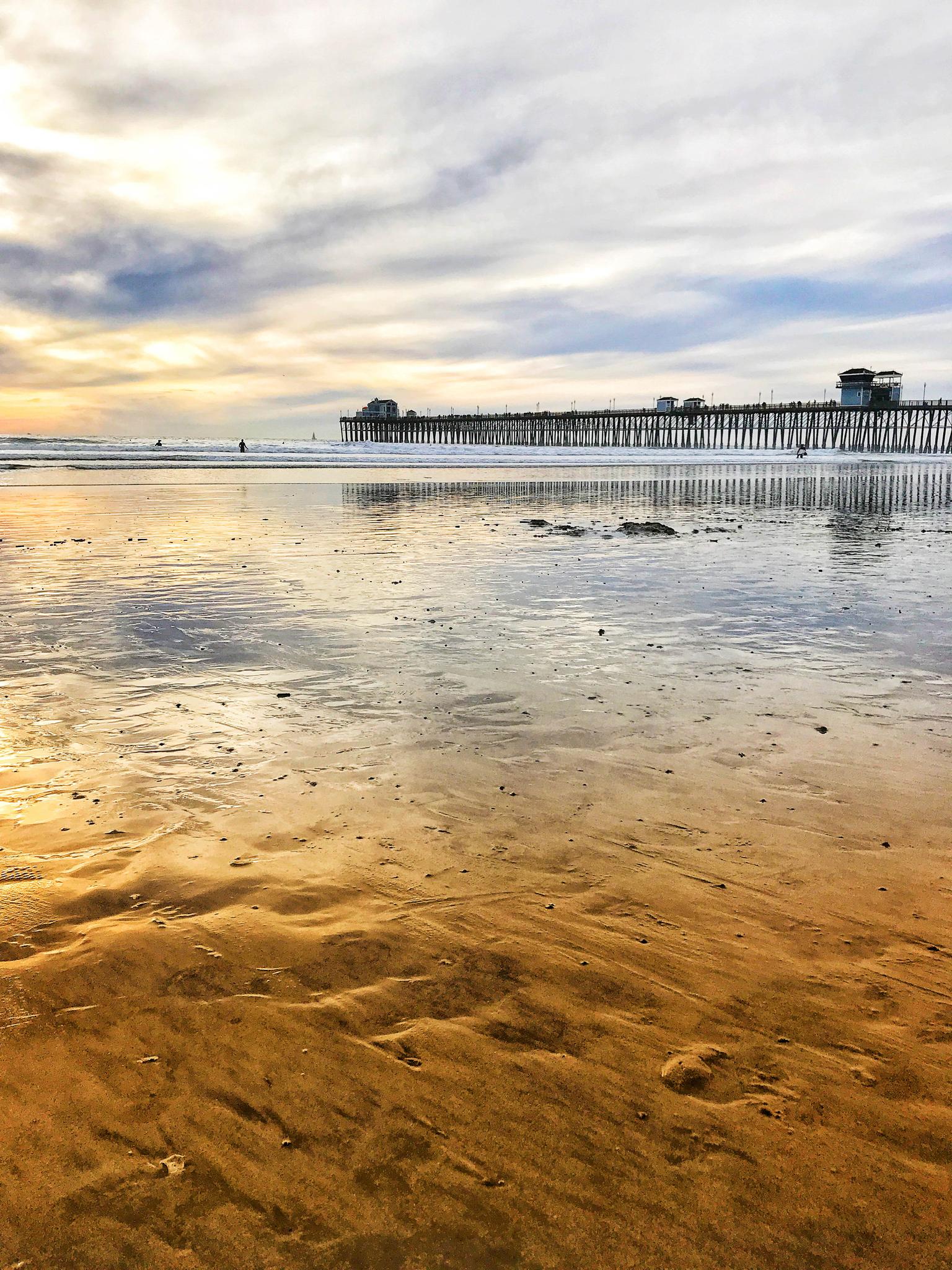 Long pier extending into the ocean at sunset, reflecting on wet sand.