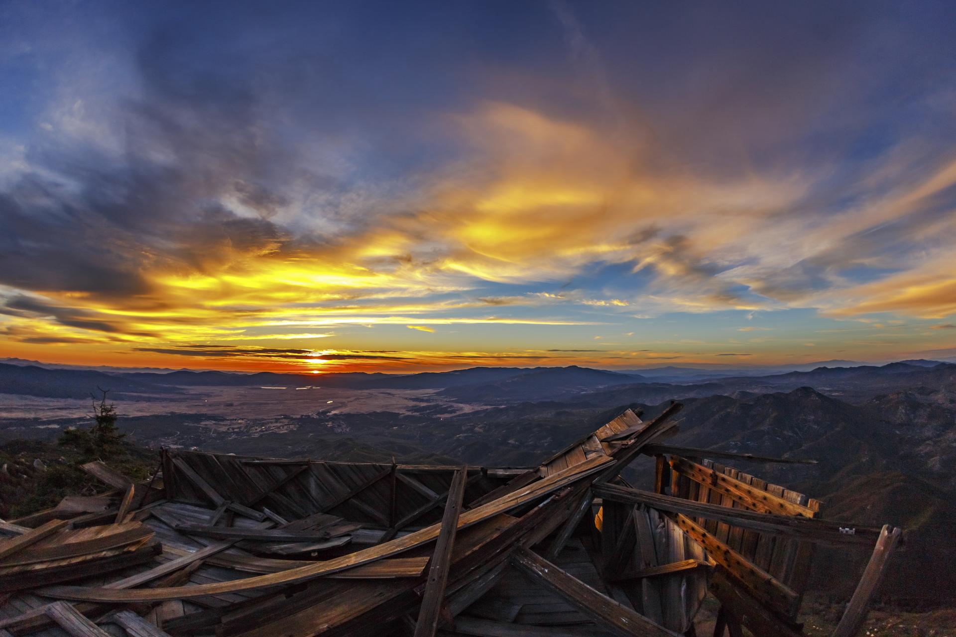 A ruined wooden structure overlooks a mountain valley under a colorful sunset sky.