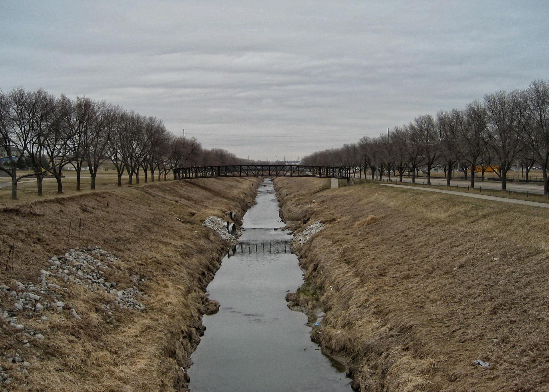 A canal with a bridge, lined by bare trees under an overcast sky.