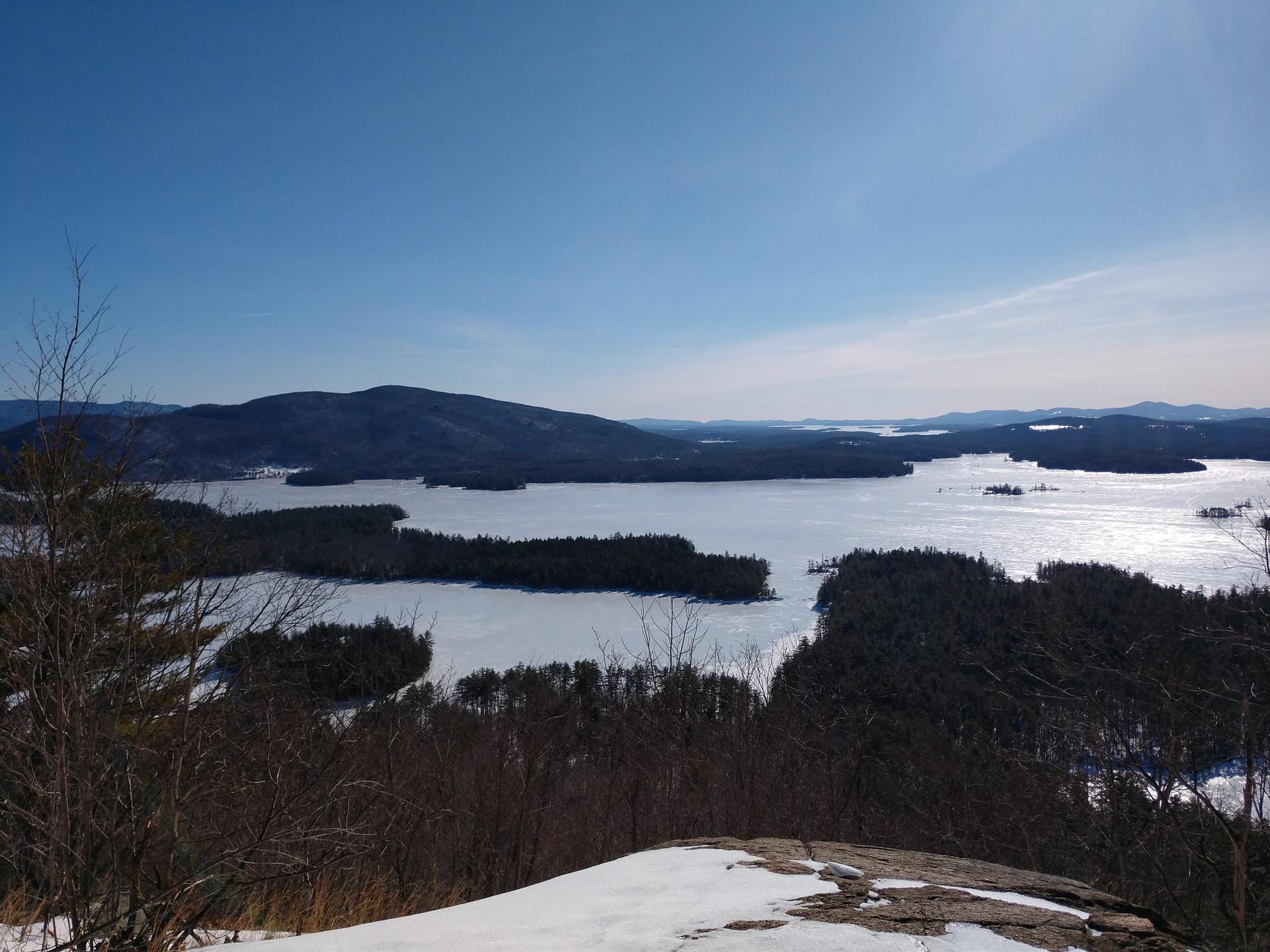 Panoramic view of a frozen lake surrounded by dark forests and distant mountains.