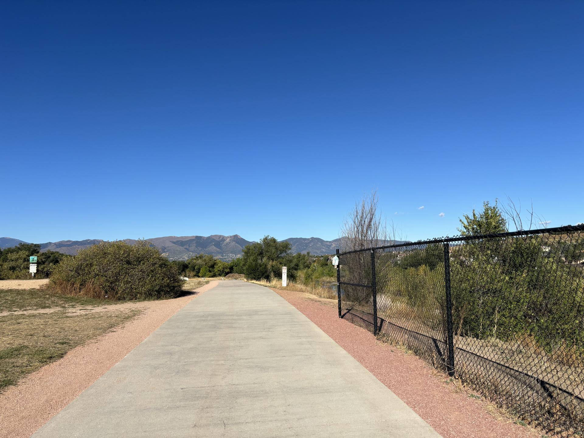 Sunny paved path with a fence and distant mountains under a clear blue sky.