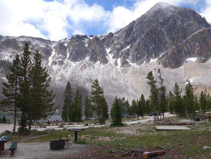 Campsite with pine trees, a lake, and snow-dusted mountains under a bright sky.