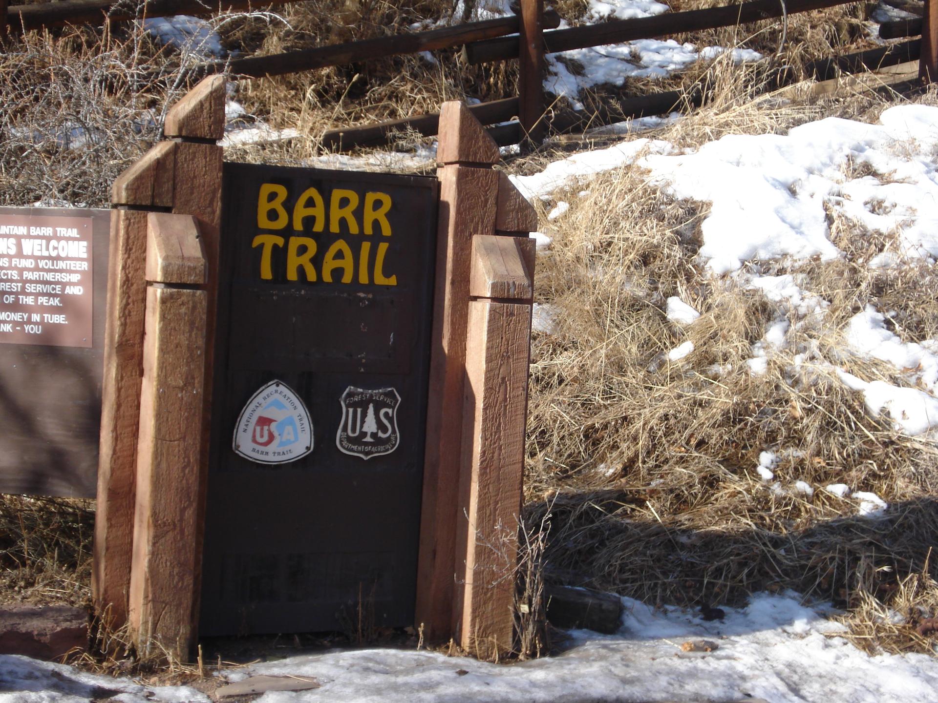 Wooden trail sign with yellow text, surrounded by dry grass and snow patches.