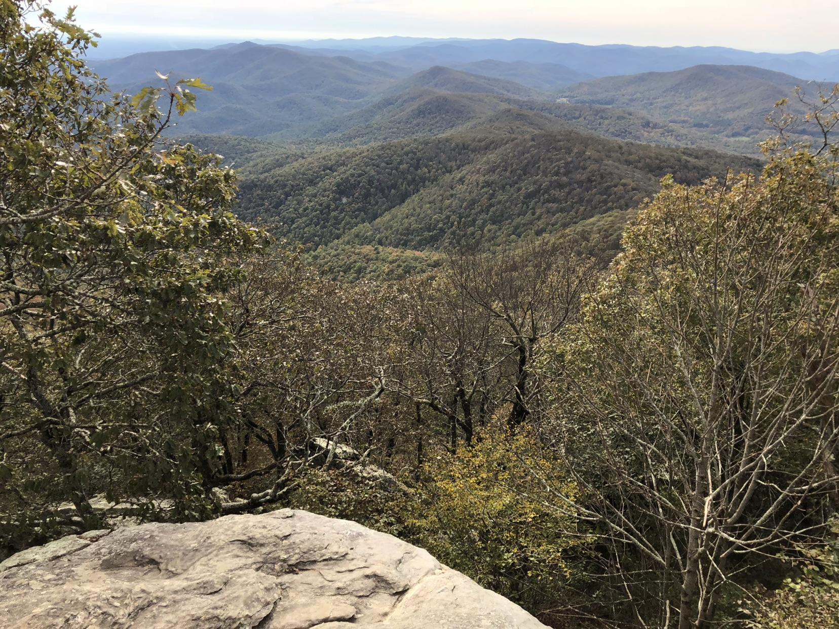 Overlook of forested mountains under a hazy sky, with rocks and trees.