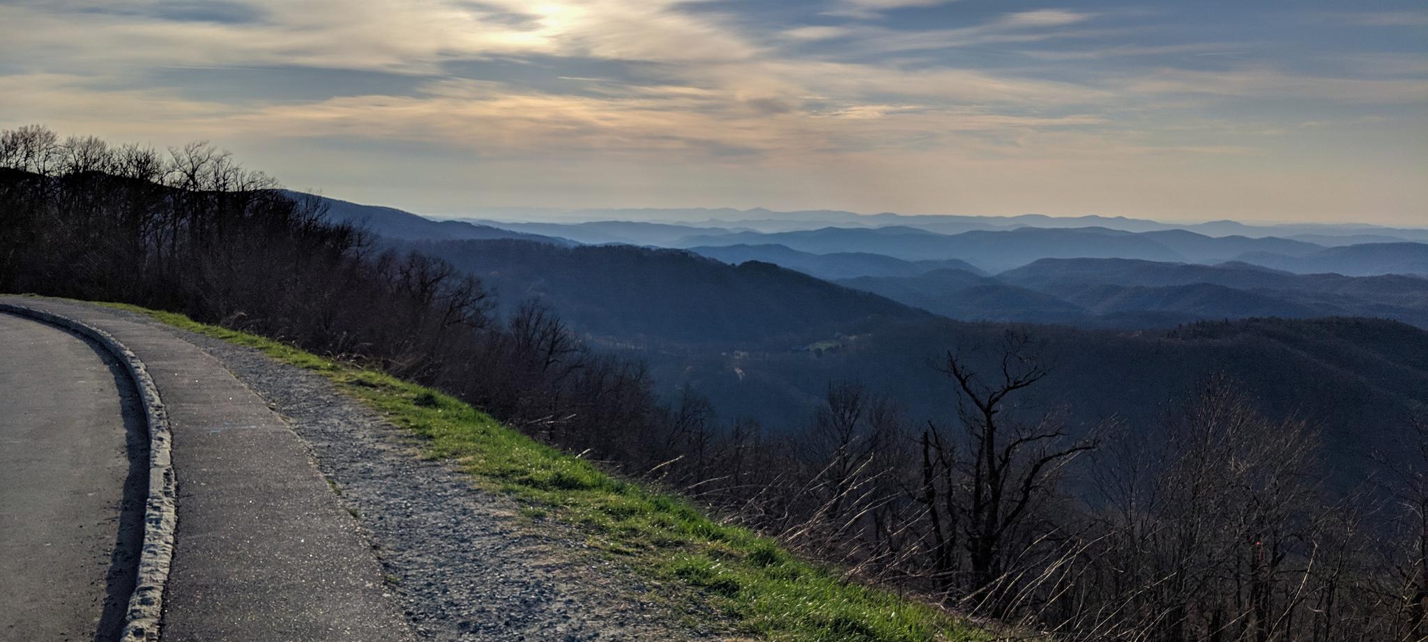 Paved road curves along a mountain ridge overlooking hazy distant peaks.