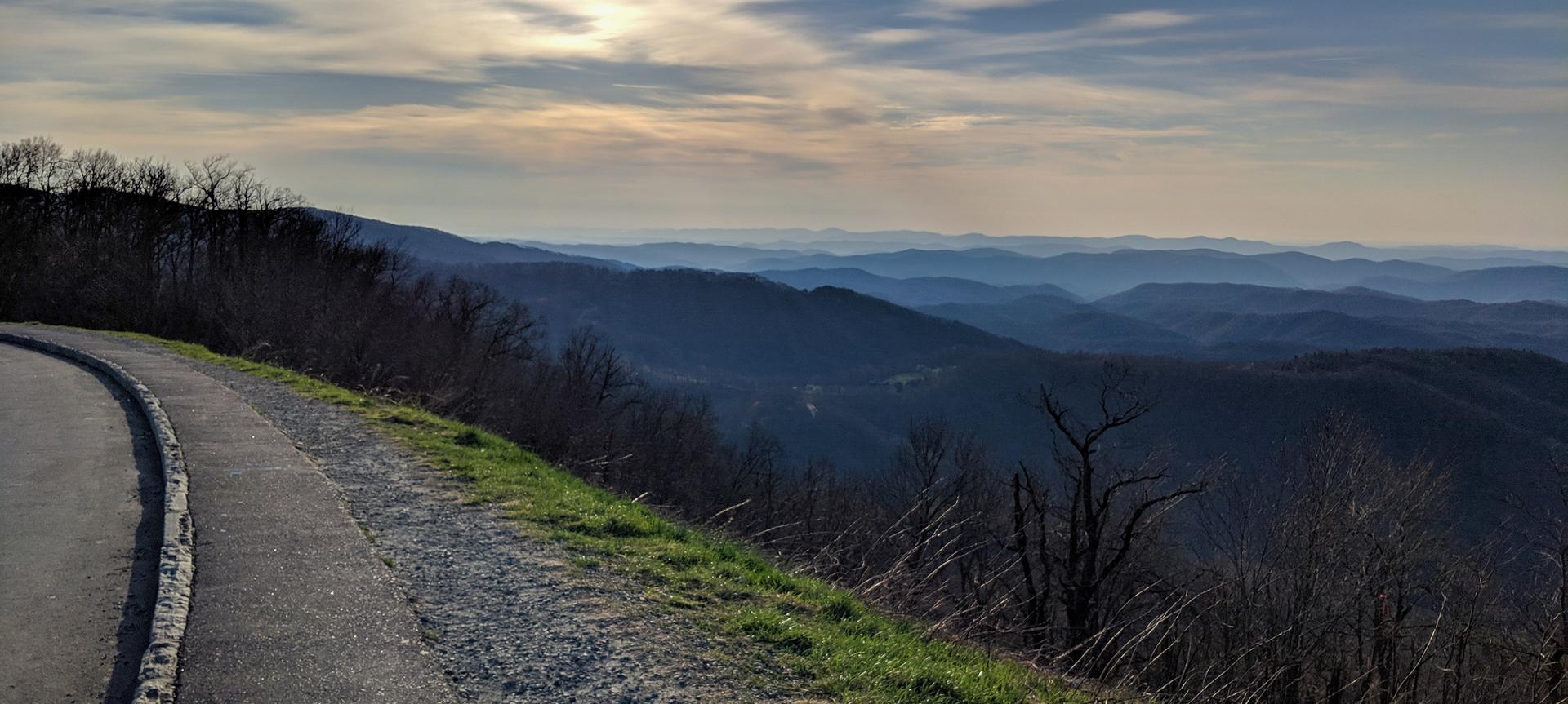 Paved road curves along a mountain ridge overlooking hazy distant peaks.