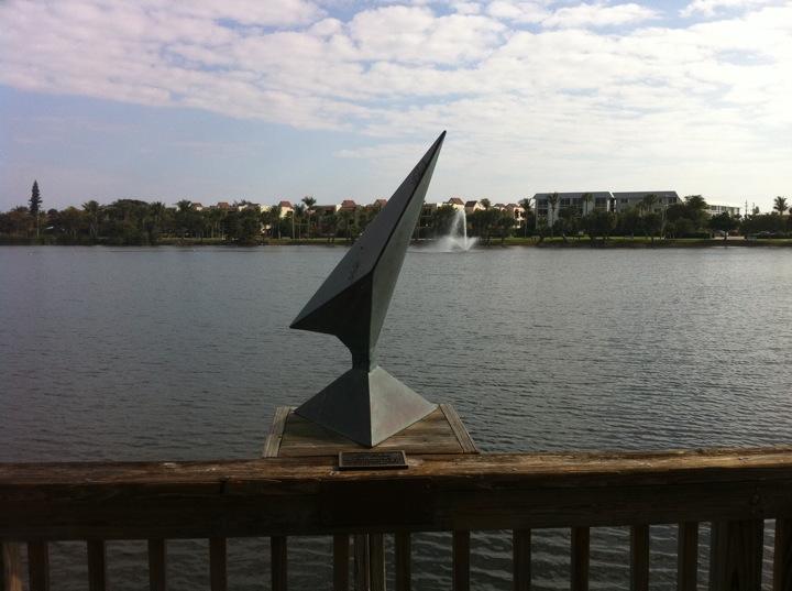 A geometric sundial sculpture on a wooden railing overlooking a lake with a fountain.