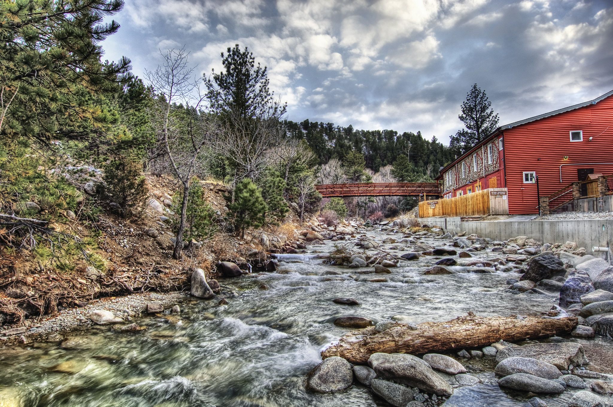 Rocky river flows past a red building and wooden bridge under cloudy sky. - Mount Princeton Hot Springs Resort and the Chalk Cliffs from Nathrop