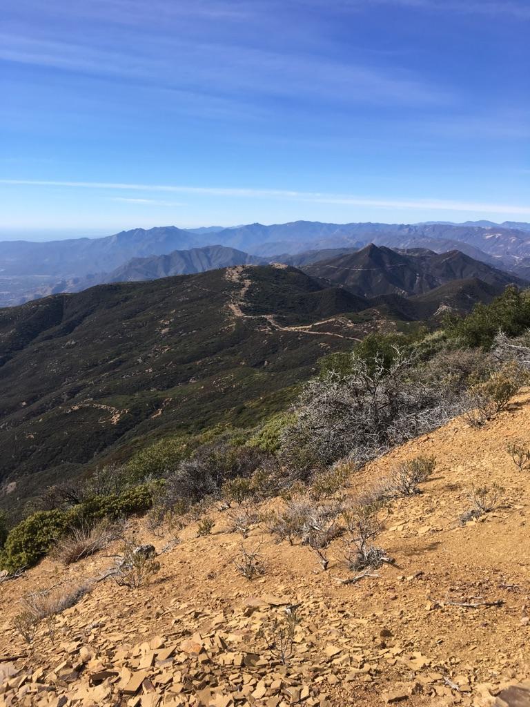 Rocky mountain slope overlooking vast, rugged green peaks under a clear blue sky.
