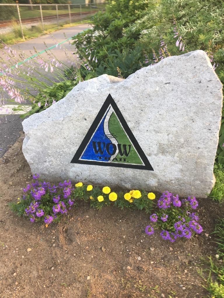Large rock with a triangular logo, surrounded by purple and yellow flowers in bright light. - Lake Winnisquam Scenic Trail – WOW Trail (Winnisquam segment) loop from Lakeport