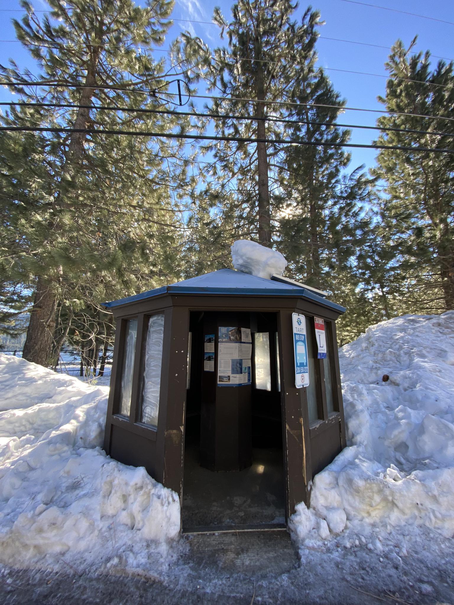 A small brown wooden shelter surrounded by deep snow and pine trees.