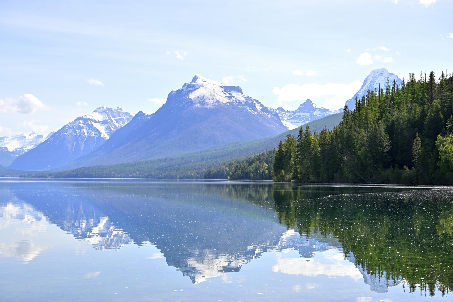 A serene lake reflects snow-capped mountains and a forested shoreline under a clear sky.