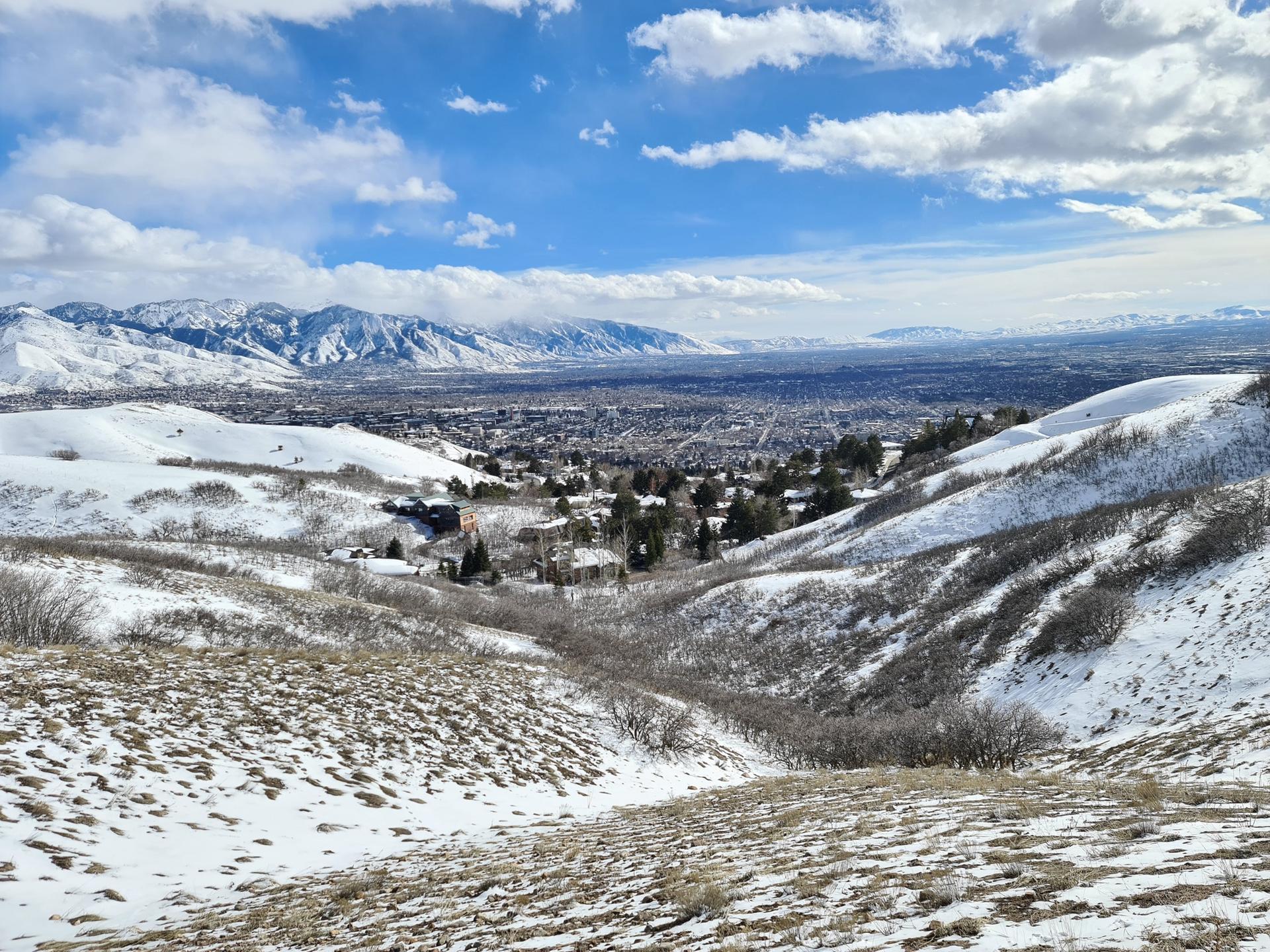 Snow-covered hills and valley with a distant city under a bright blue sky.