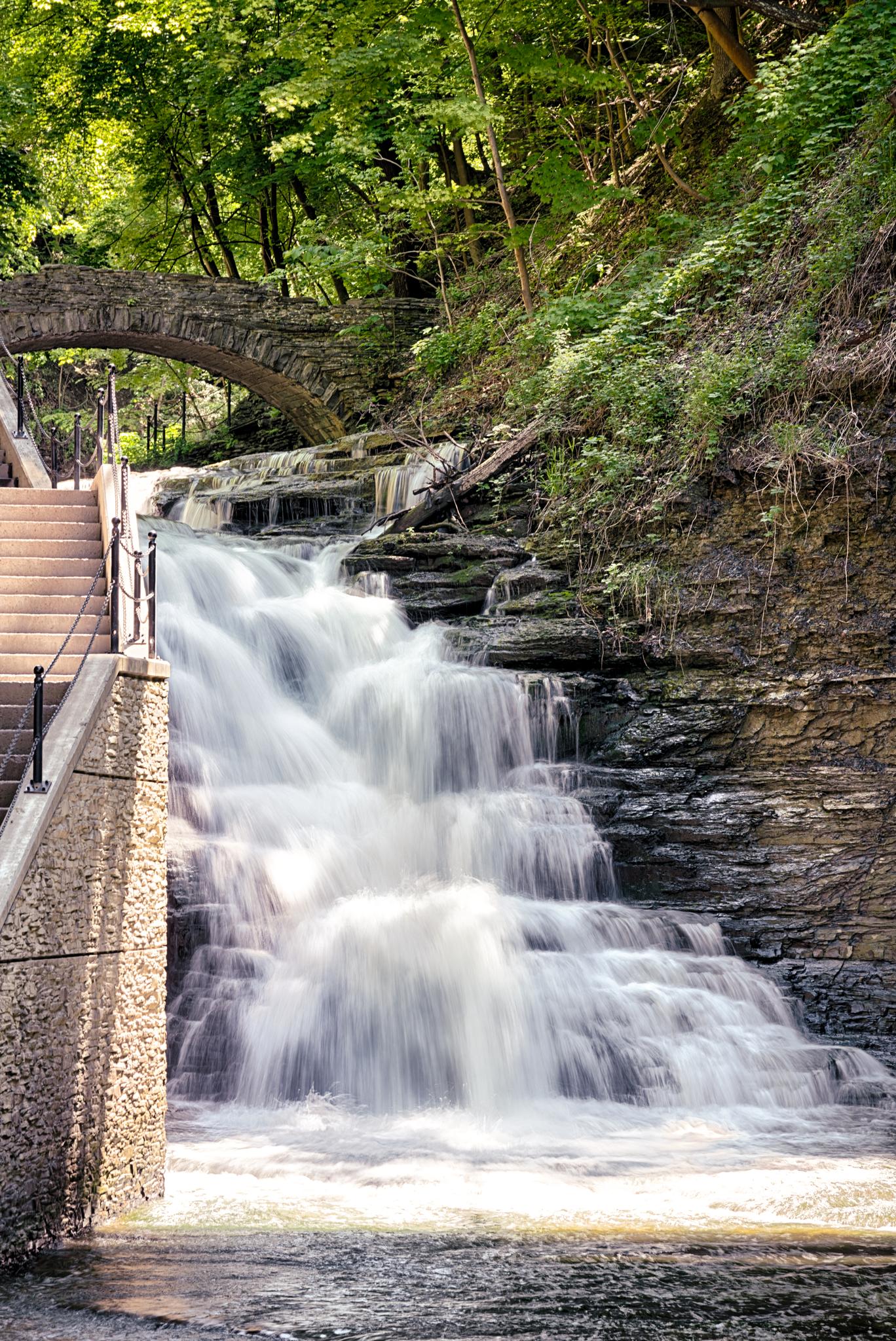 Cascading waterfall next to stone stairs and an arched bridge in a sunny gorge.