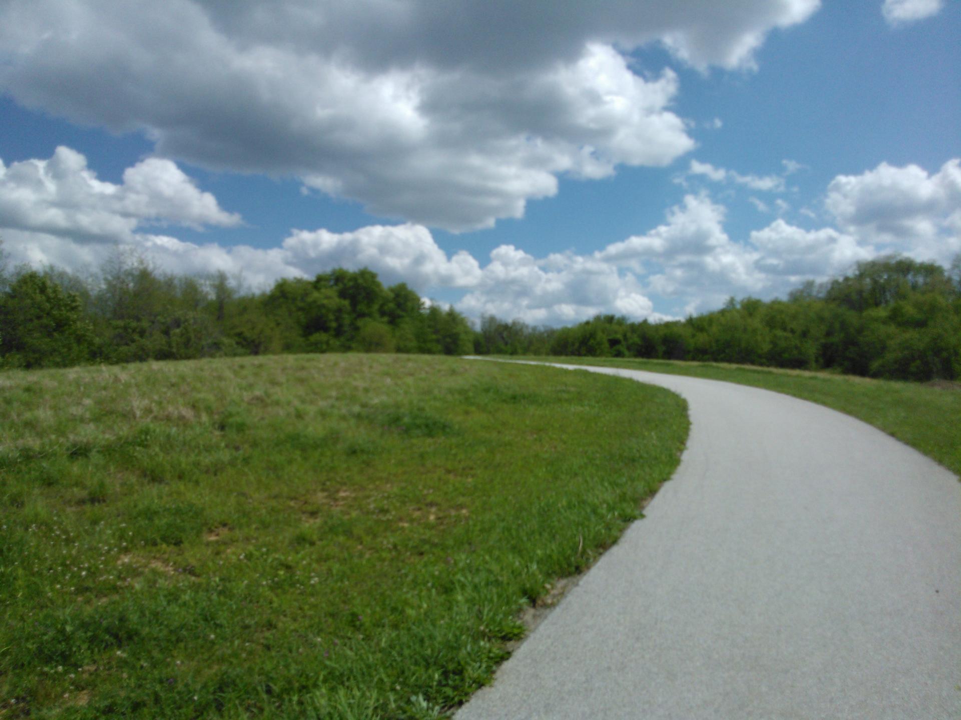 A paved path curves through a green field under a bright, cloudy sky.