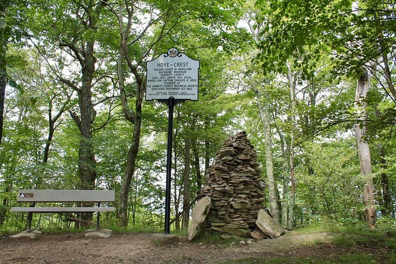 A white sign, stone cairn, and bench in a bright, green forest.