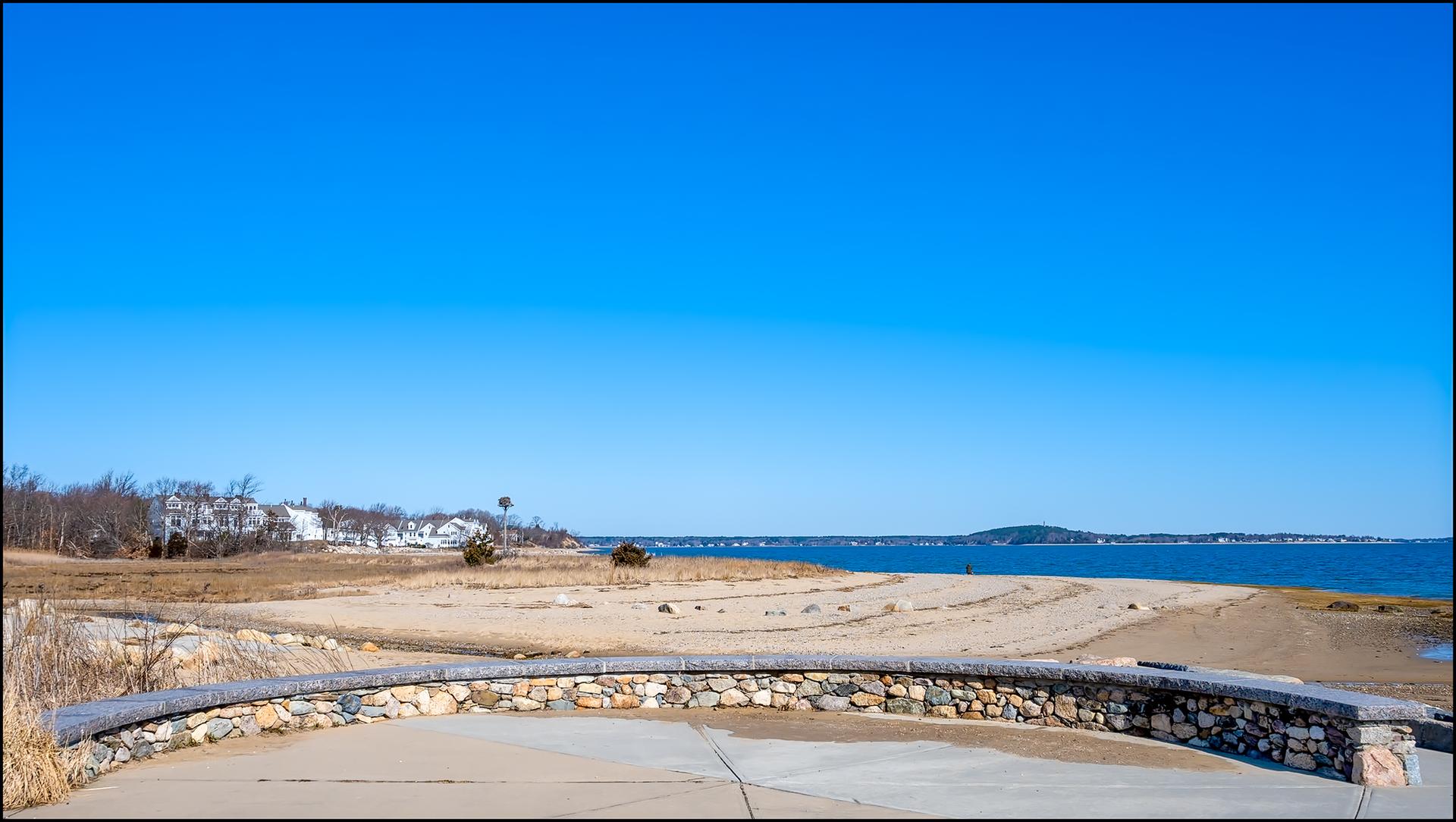 A wide sandy beach and blue ocean under a clear sky, stone wall foreground.