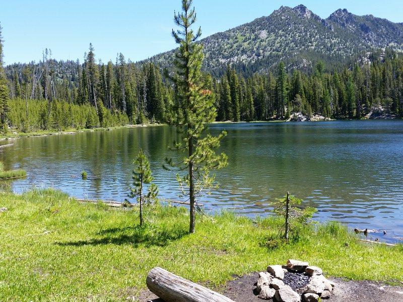 A serene lake with forested shores, mountains, and a stone fire pit under sunny skies.