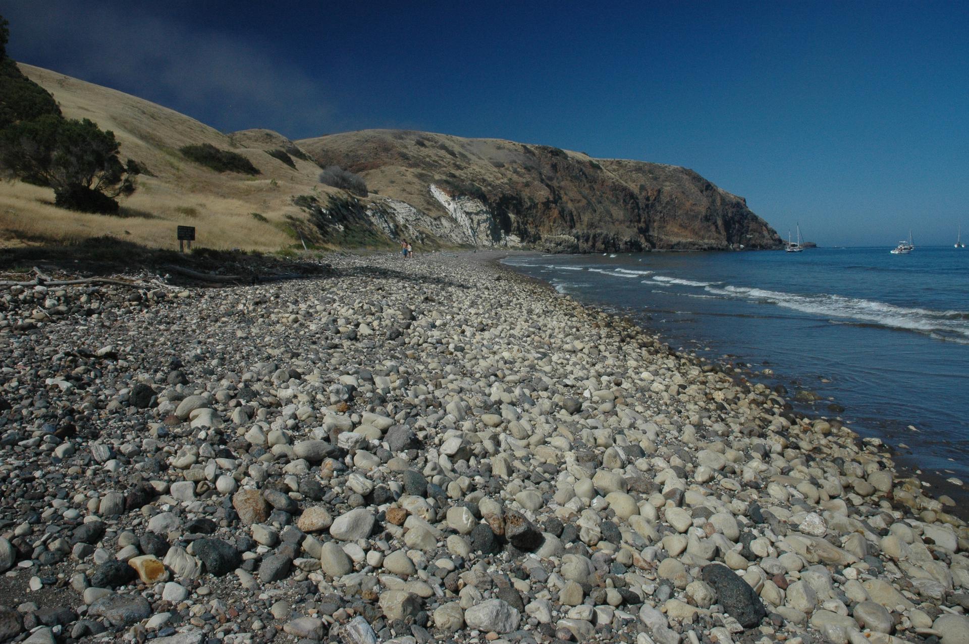 Sunny view of a wide rocky beach with waves and steep coastal cliffs.