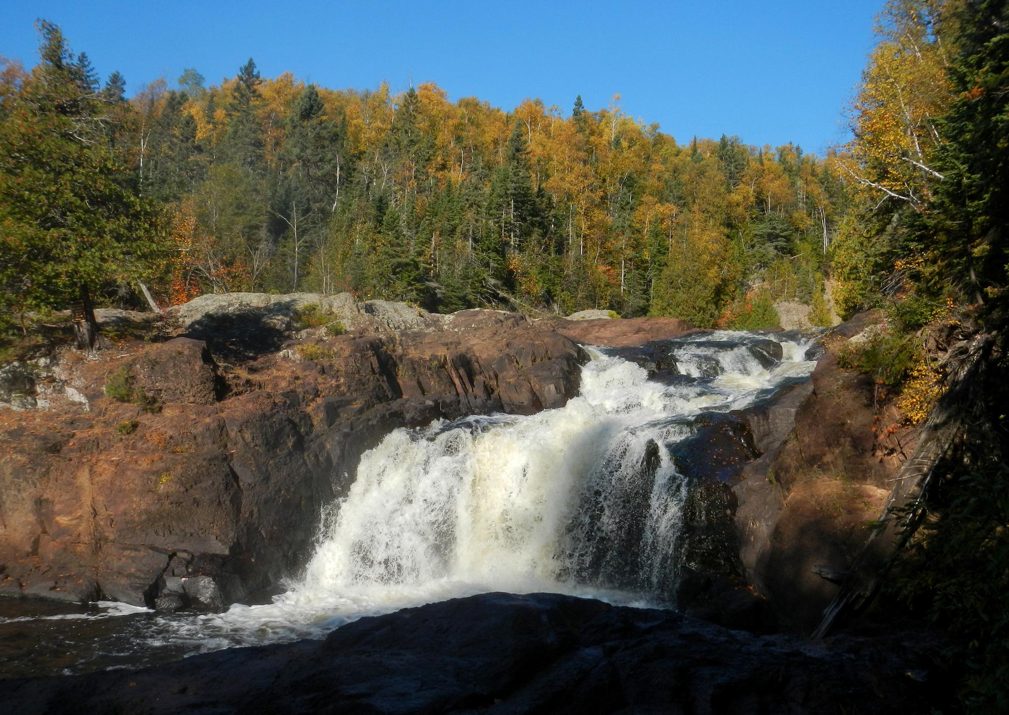 Wide waterfall cascades over dark rocks, backed by a forest with autumn colors.