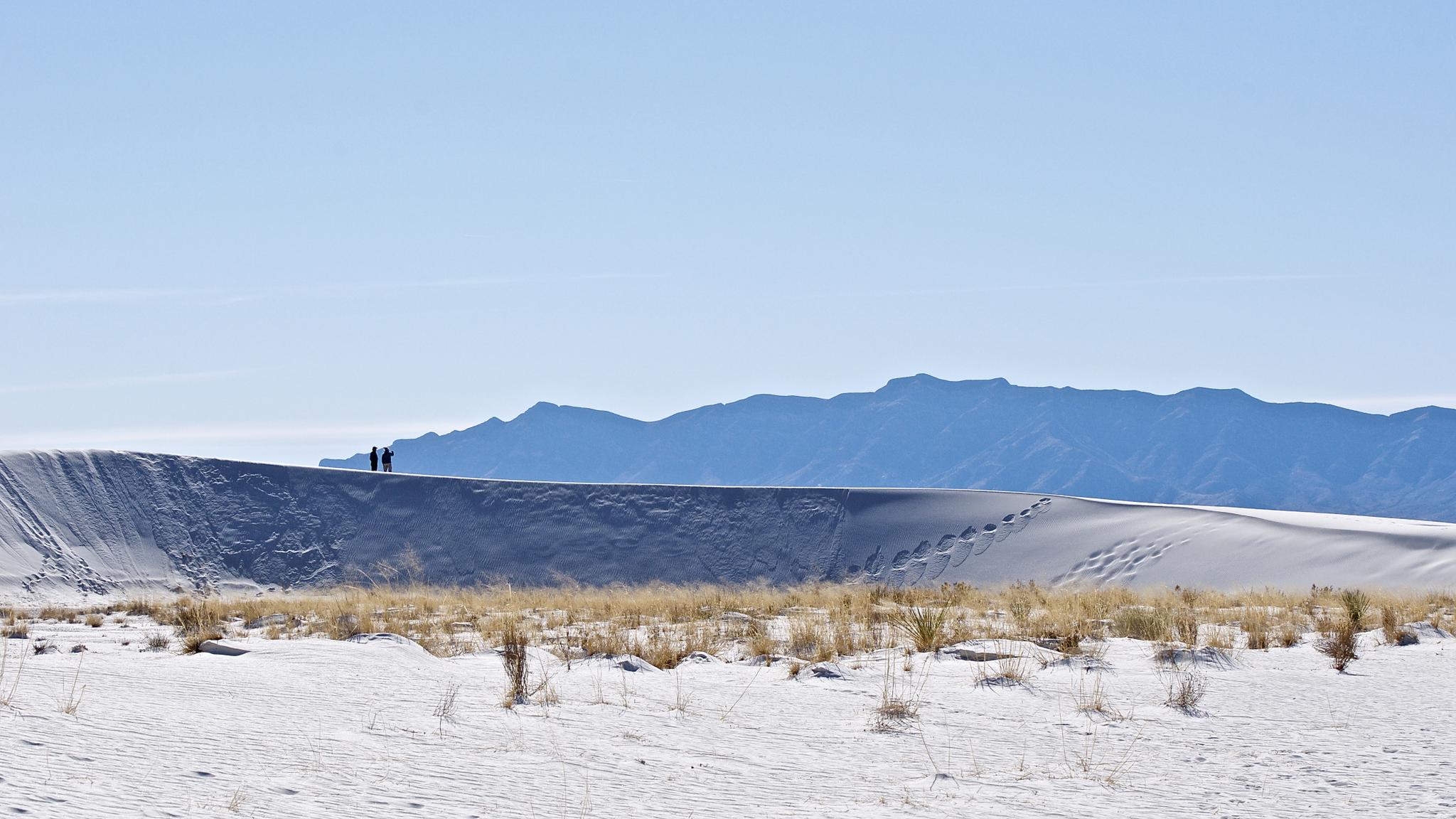 Two people stand atop a white sand dune with distant mountains.