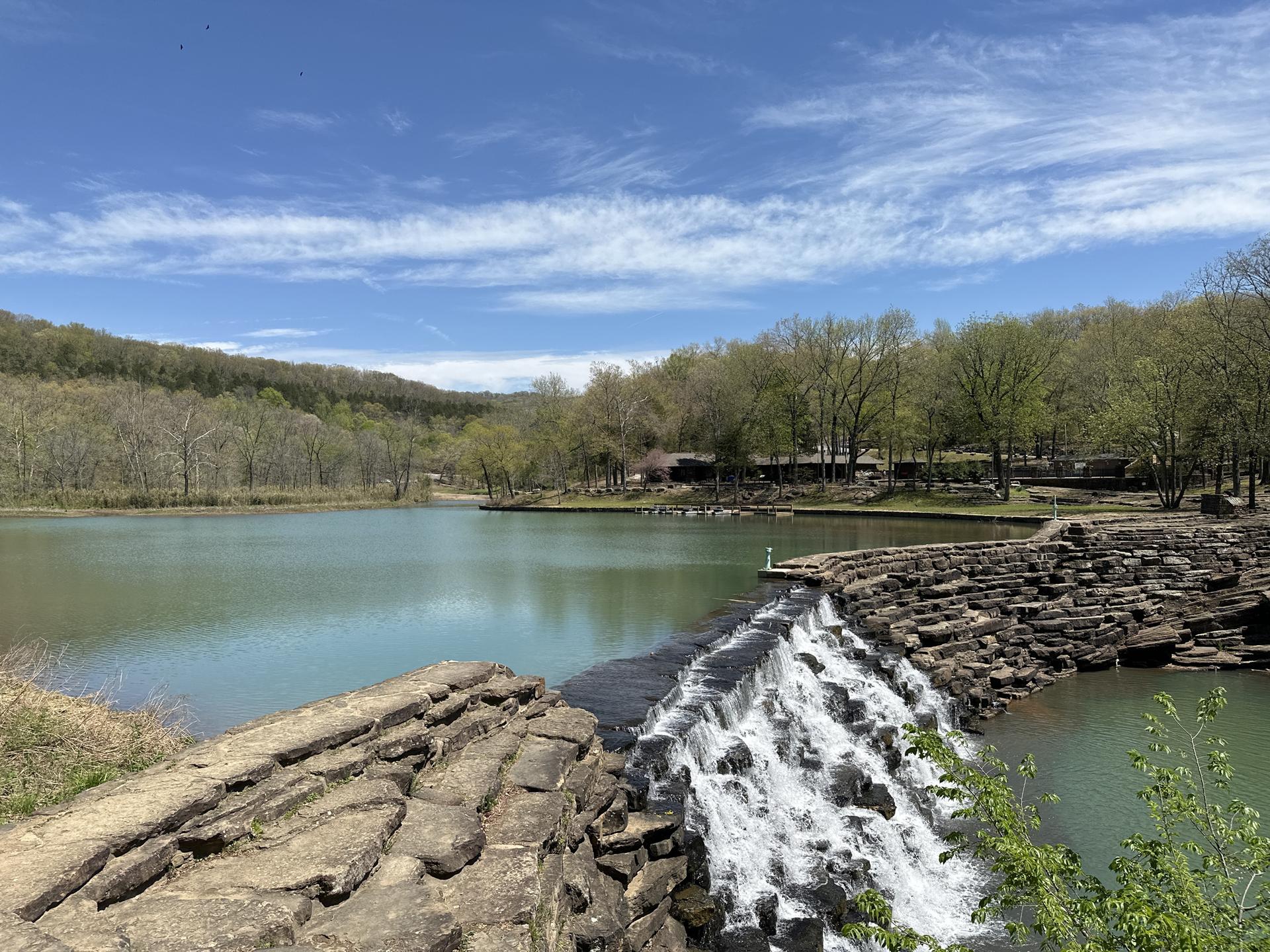 Sunny day over a lake with a rocky waterfall and forested hills.