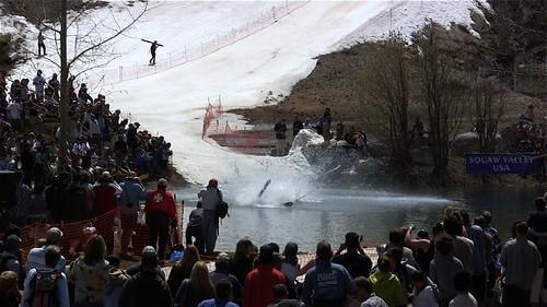 Crowd watches a skier splash into a pond from a snow ramp on a sunny day.