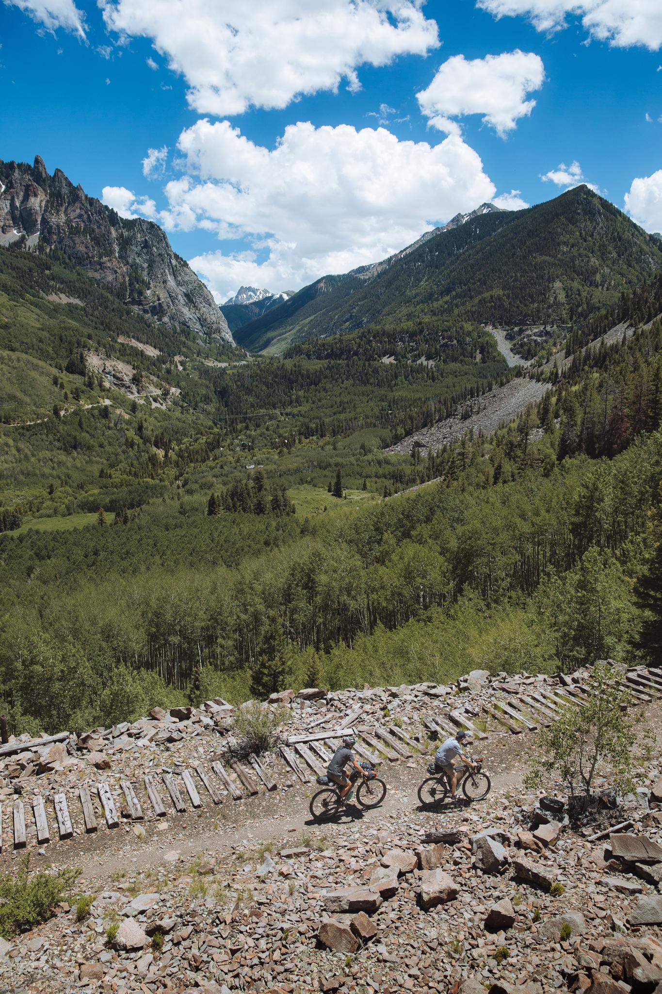 Two mountain bikers on a rocky trail overlooking a lush, forested valley.