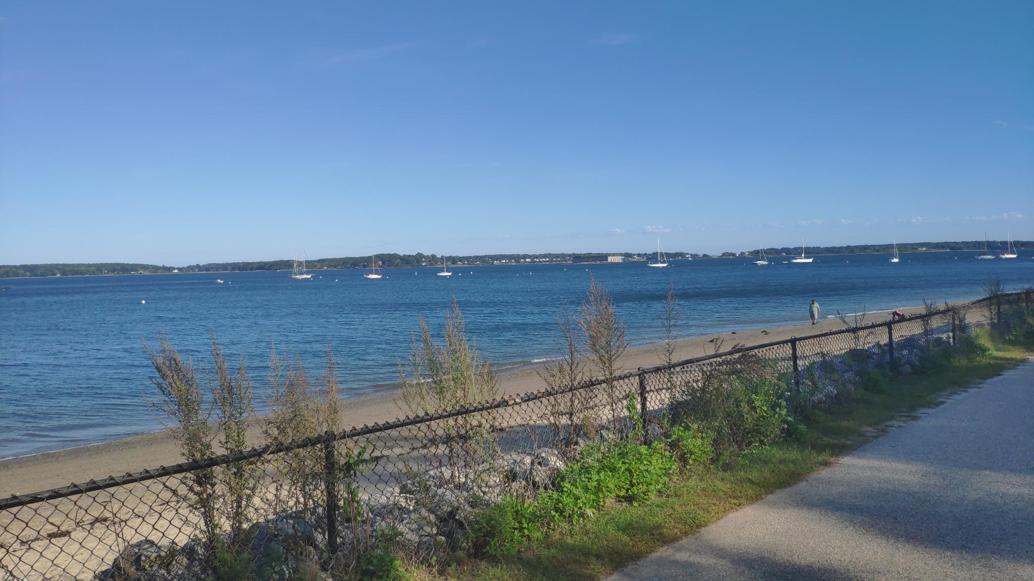 Sunny beach with calm water, distant boats, and a fence in the foreground.