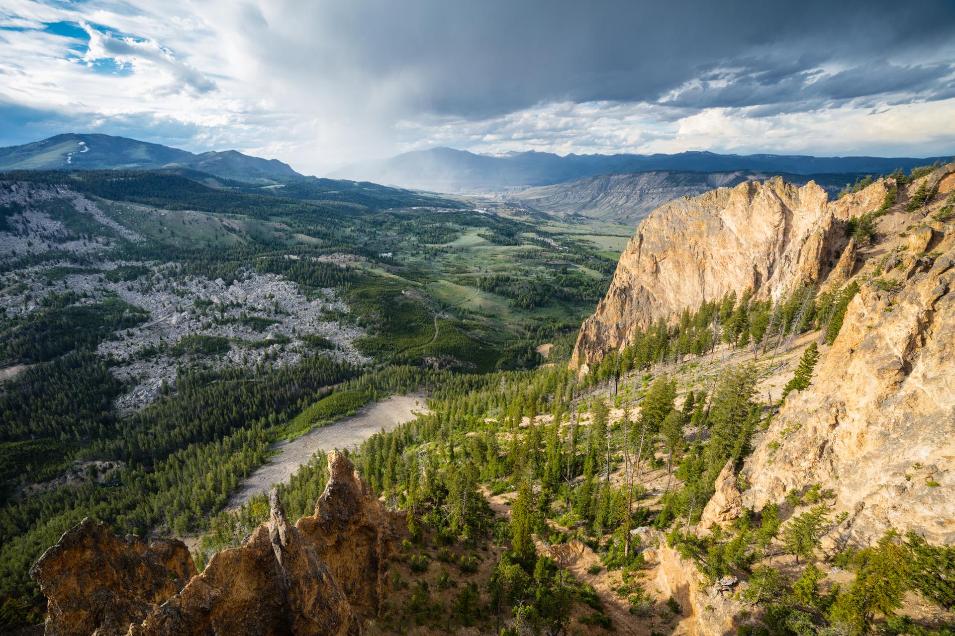 Dramatic mountain landscape with steep cliffs, a forested valley, and stormy skies.