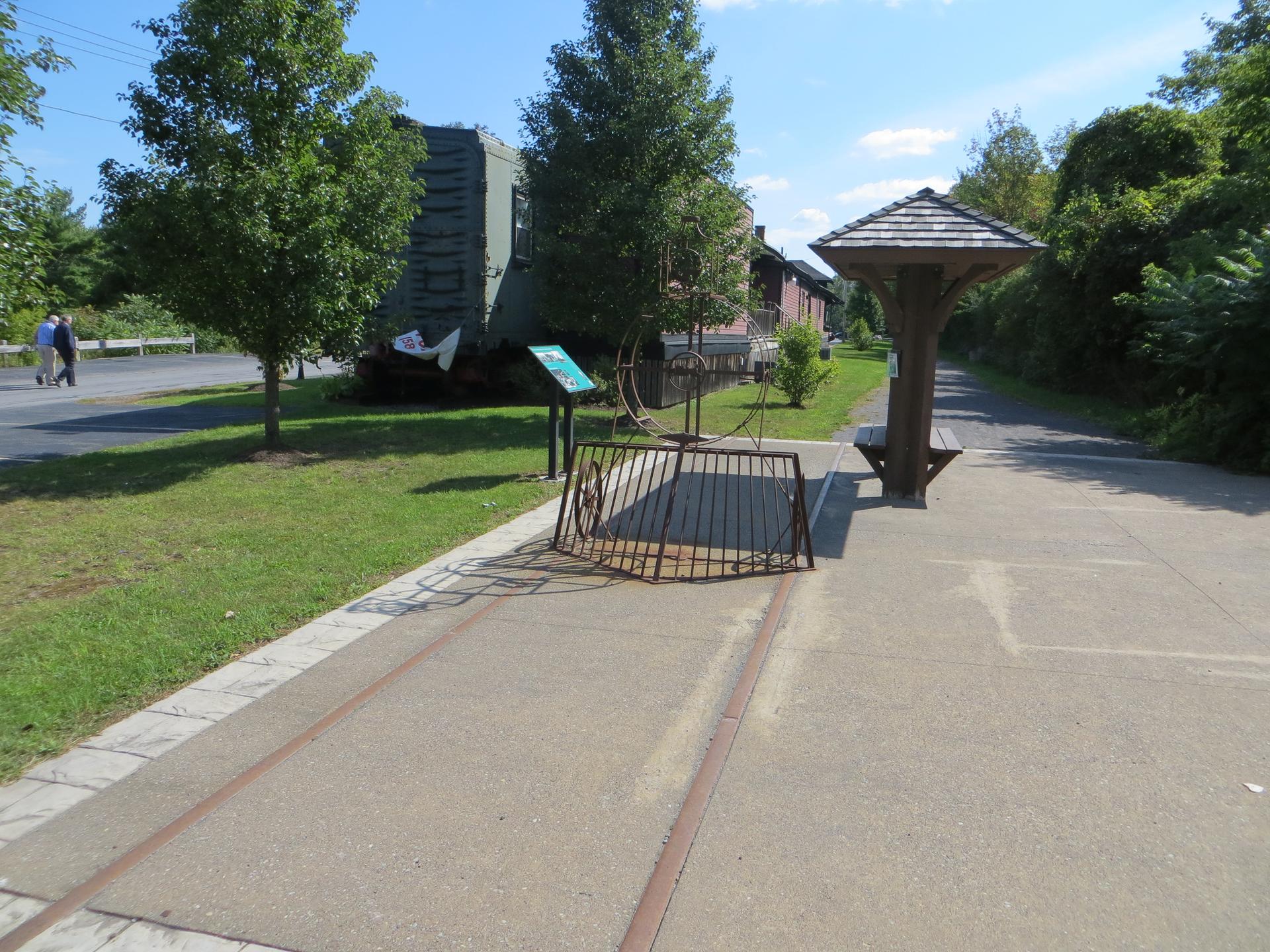 Paved path with embedded tracks, a shelter, and distant walkers on a sunny day.