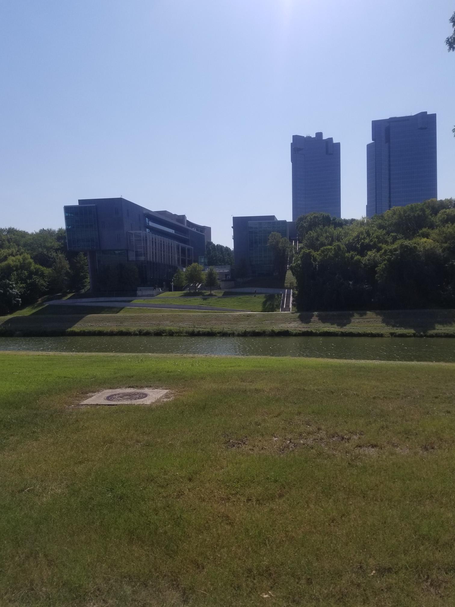 Sunny view of a river, green banks, and tall city buildings in the distance.