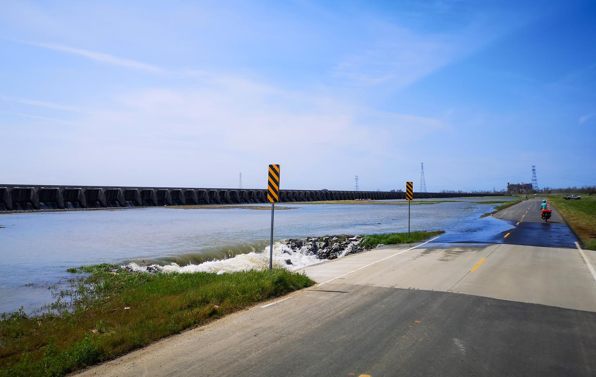 A road next to a wide body of water with a distant cyclist under blue sky.