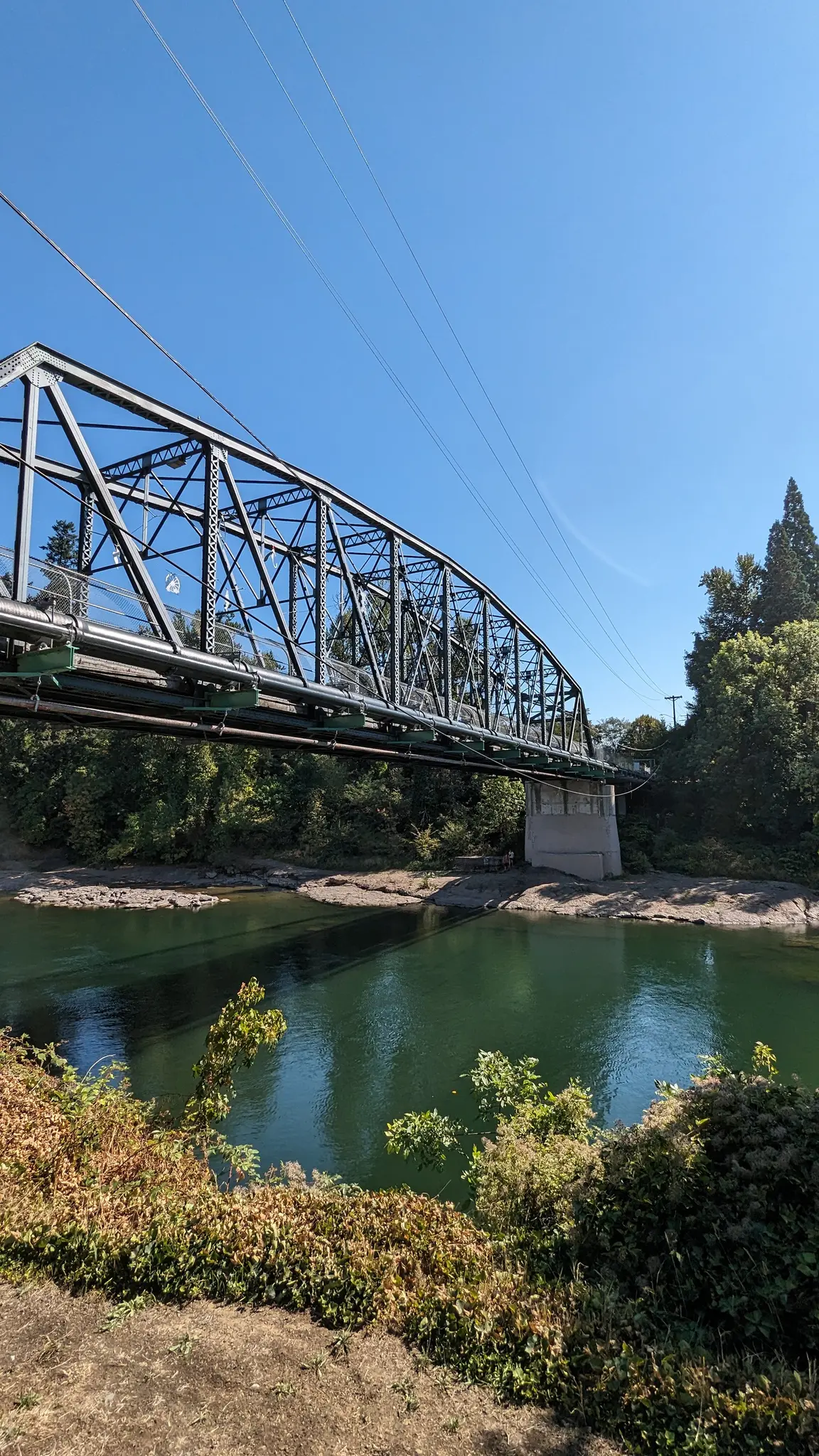 A large metal truss bridge spans a green river under a clear blue sky.
