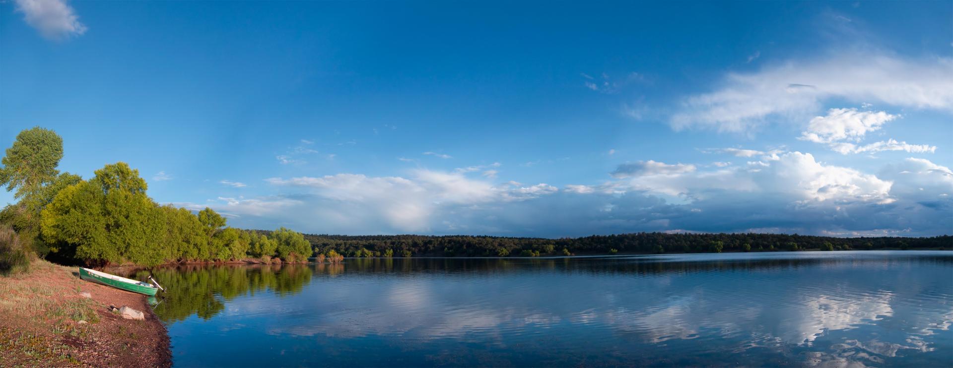 Calm lake reflecting clouds, with a green boat on the tree-lined shore.