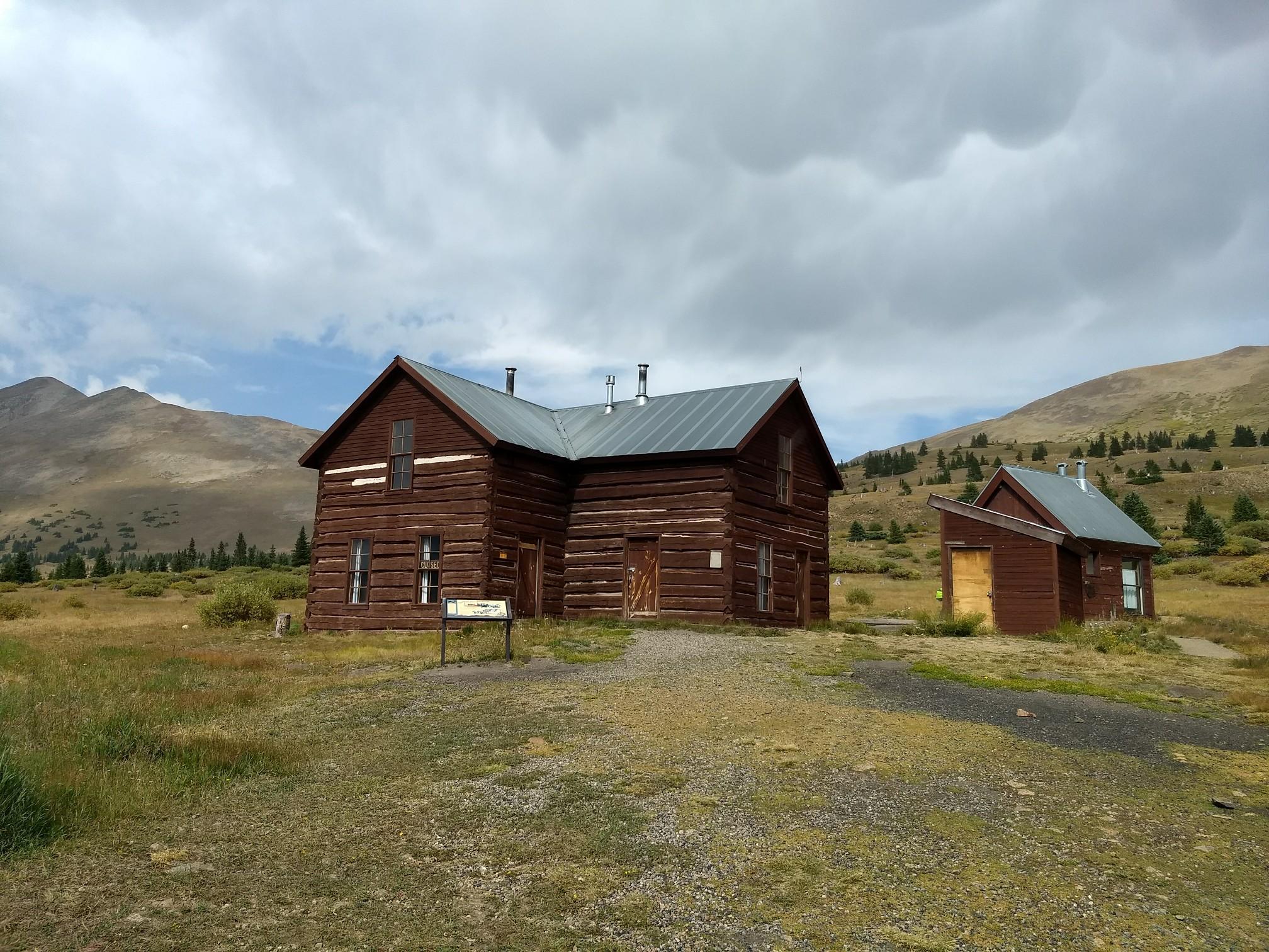 Two rustic wooden cabins stand on a grassy hillside under a cloudy sky.