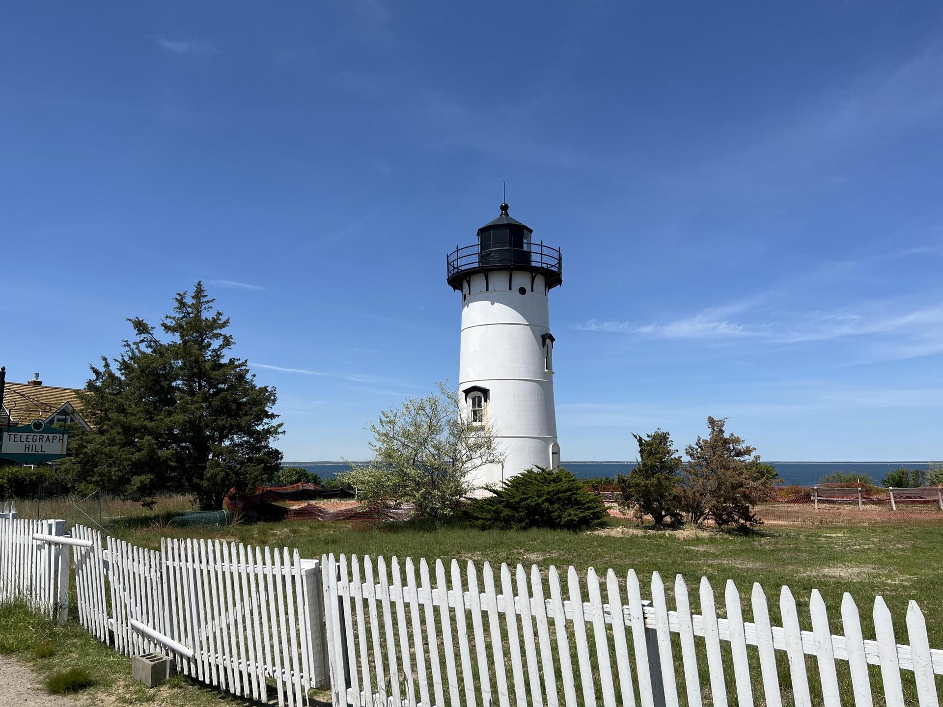 White lighthouse with a black top behind a white picket fence.