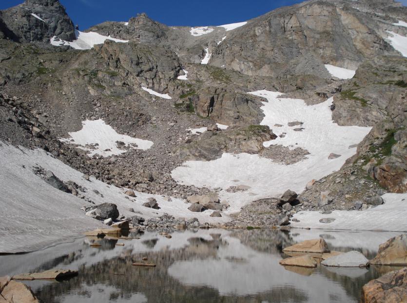 Alpine lake reflecting snowy, rocky mountains under a bright blue sky.