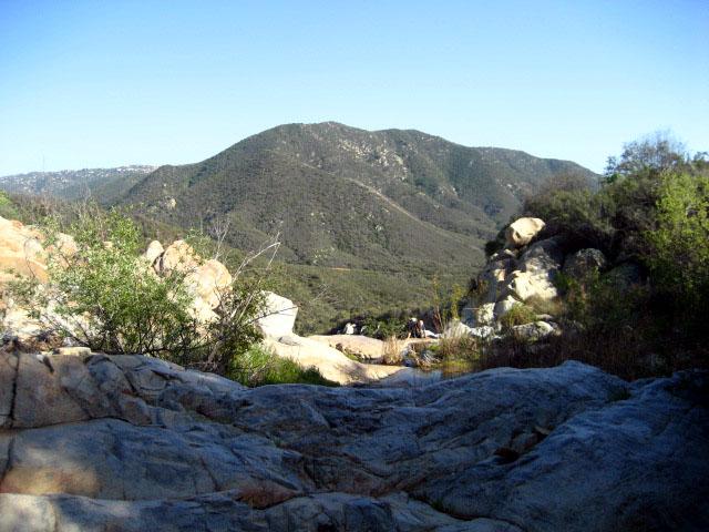 Sunny mountain view with rocky foreground, green slopes, and small figures hiking.