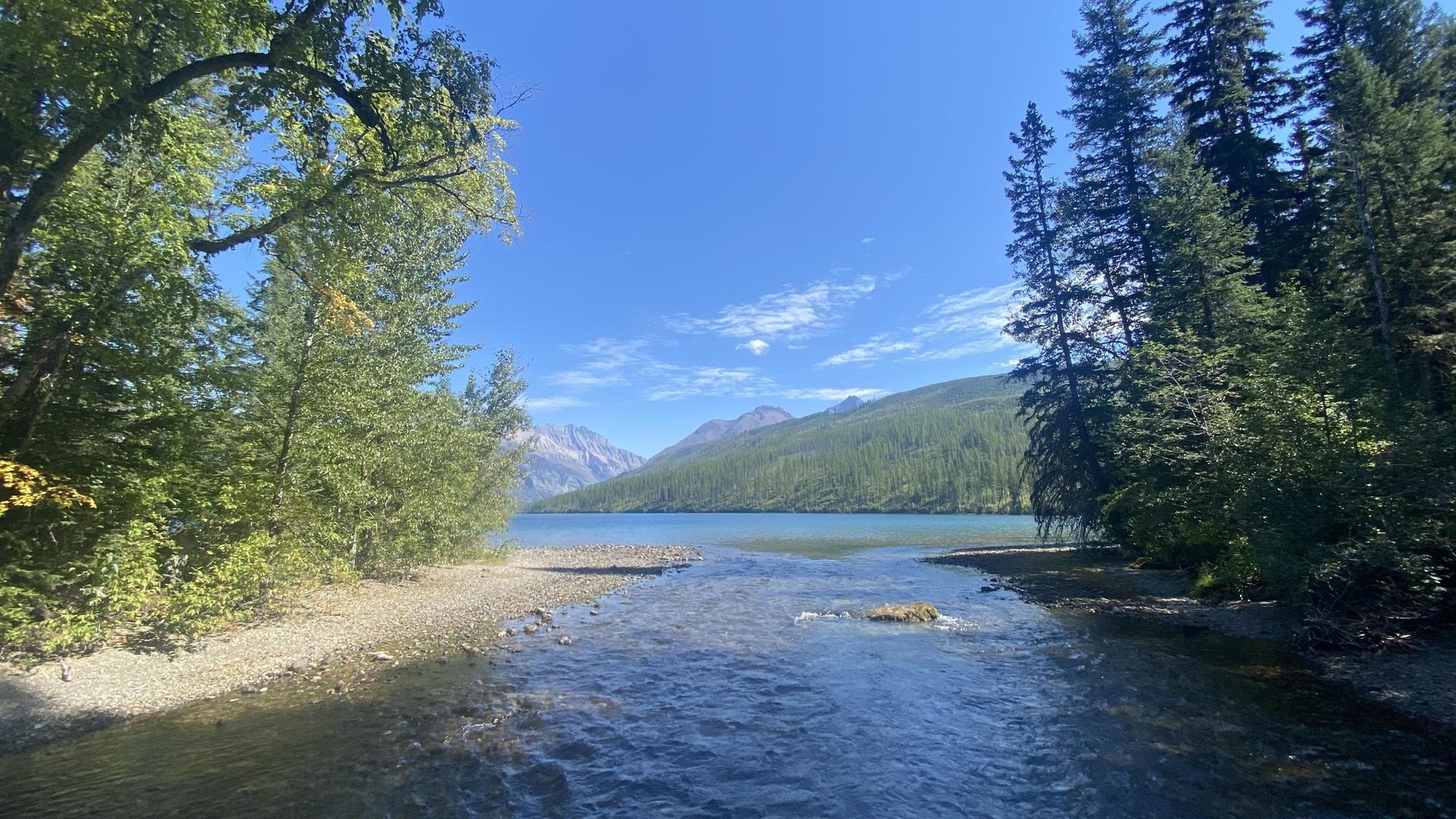 Stream flows into a wide lake with distant mountains under a blue sky.
